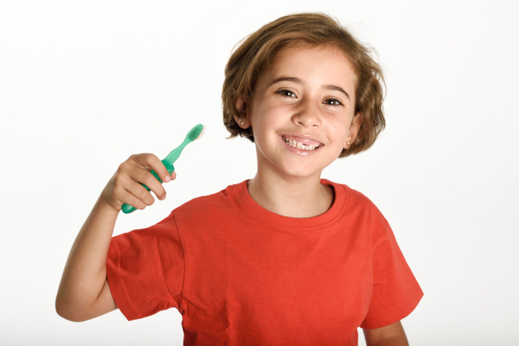 Criança de blusa vermelha e uma escova de dentes na mão para falar sobre a ação do flúor na prevenção de cáries.