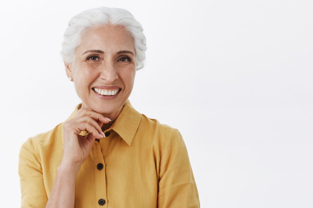Mulher acima dos 60 anos com blusa amarela, sorrindo e com a mão abaixo do queixo, a fim de falar sobre a síndrome do envelhecimento bucal precoce.