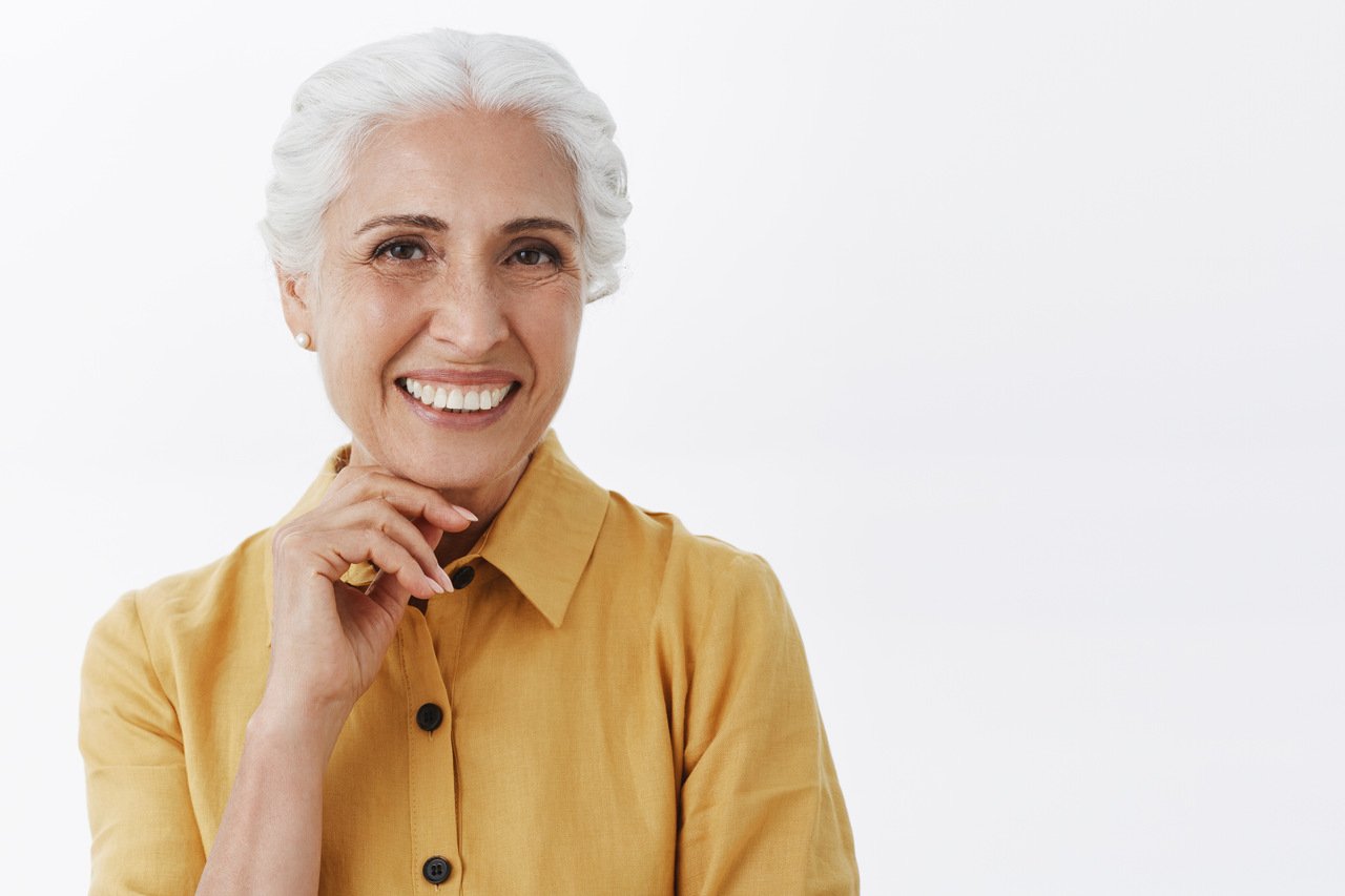 Mulher acima dos 60 anos com blusa amarela, sorrindo e com a mão abaixo do queixo, a fim de falar sobre a síndrome do envelhecimento bucal precoce.